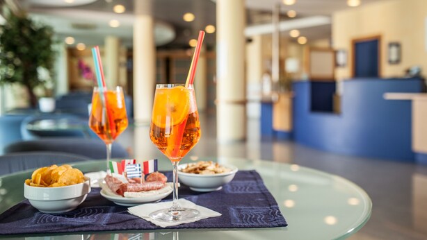 Una mesa con un vaso de zumo de naranja, un vaso de vino tinto, un bol de patatas fritas y un plato de albóndigas, sobre una mesa de cristal en el vestíbulo de un hotel.