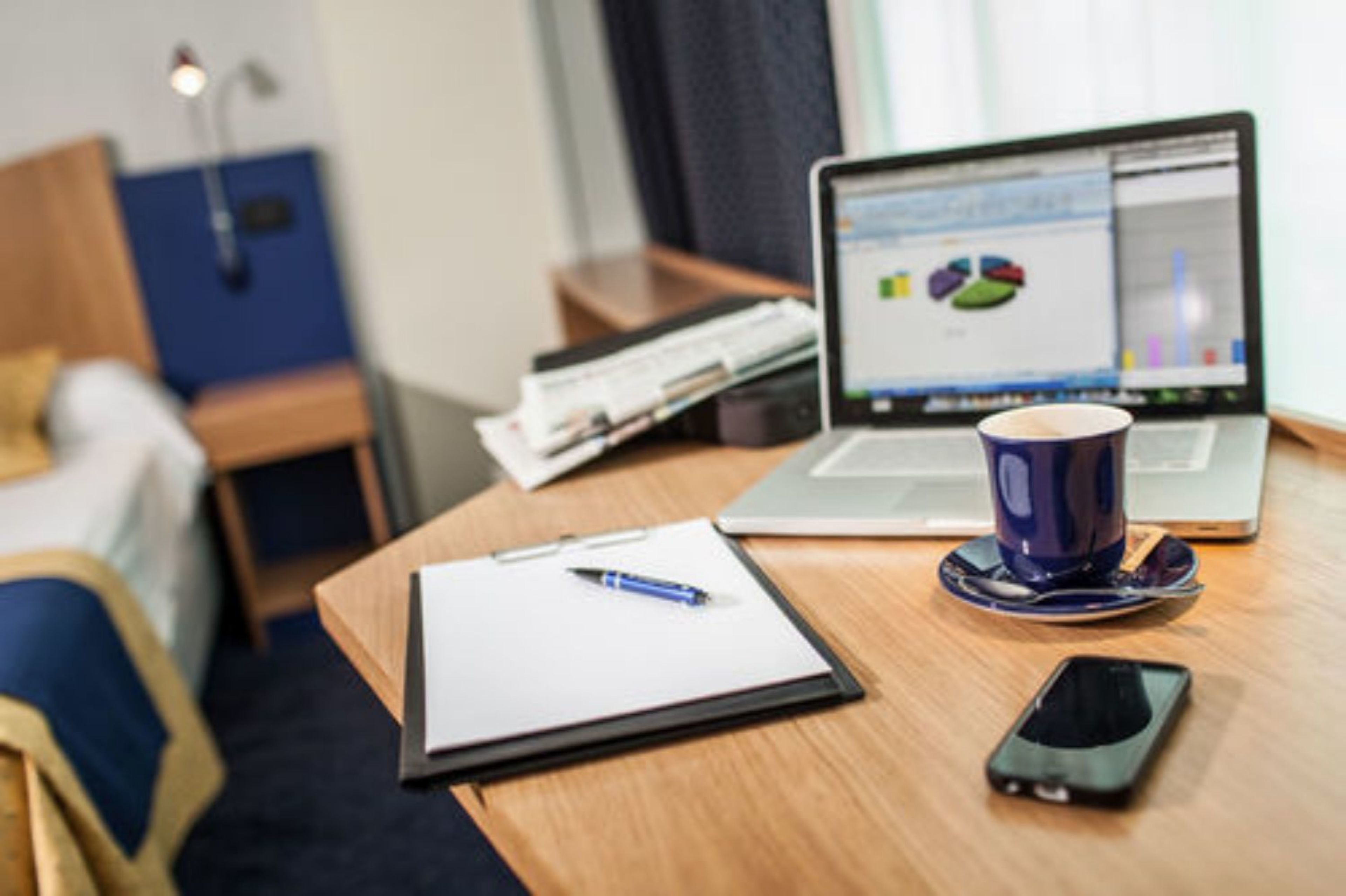Une chambre d’hôtel avec un lit, un bureau avec un ordinateur portable, un carnet, un stylo, un téléphone intelligent et une tasse à café.