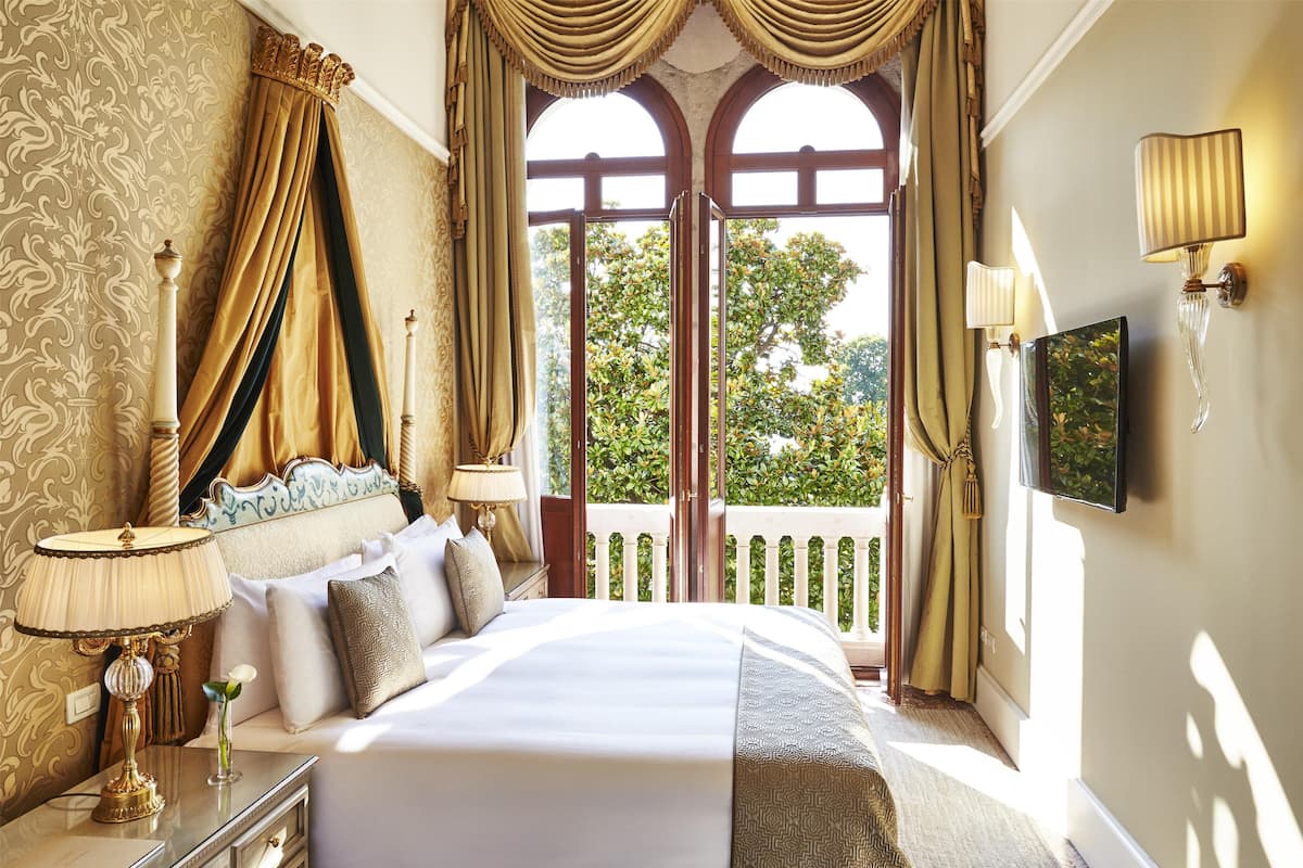 A hotel room with a large bed, curtains, a TV, and a balcony with a view of trees.