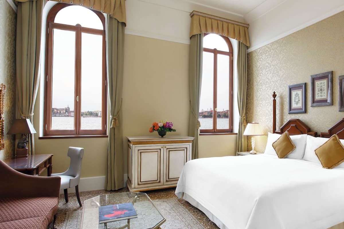 A hotel room with a large bed, a desk with a chair, a small table with a book, and two windows with curtains.