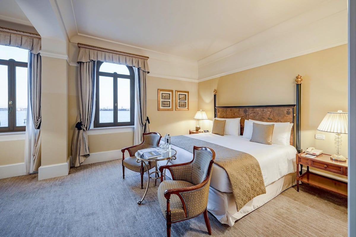 A hotel room with a large bed, two chairs, a table, a lamp, and a view of the sea through the window.