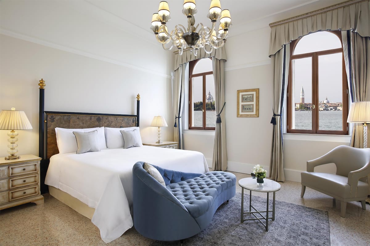 A hotel room with a large bed, a curved blue sofa, a small round table with a vase, and a view of a body of water through large windows.