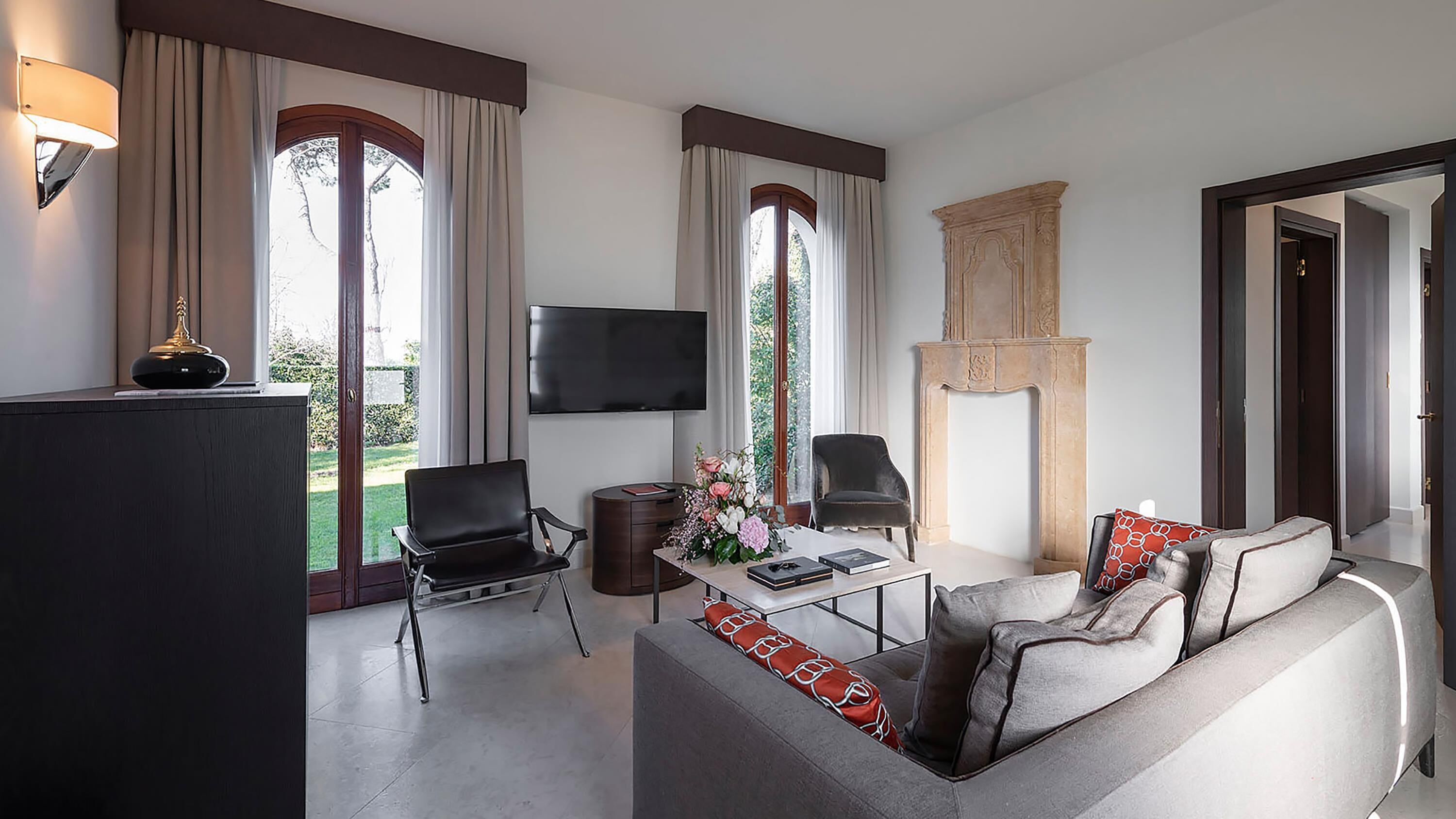 Un salon moderne comprenant un canapé, deux fauteuils, une table basse et un téléviseur à écran plat. De grandes fenêtres sont agrémentées de rideaux, et on y trouve une cheminée ainsi qu’un téléviseur fixé au mur.