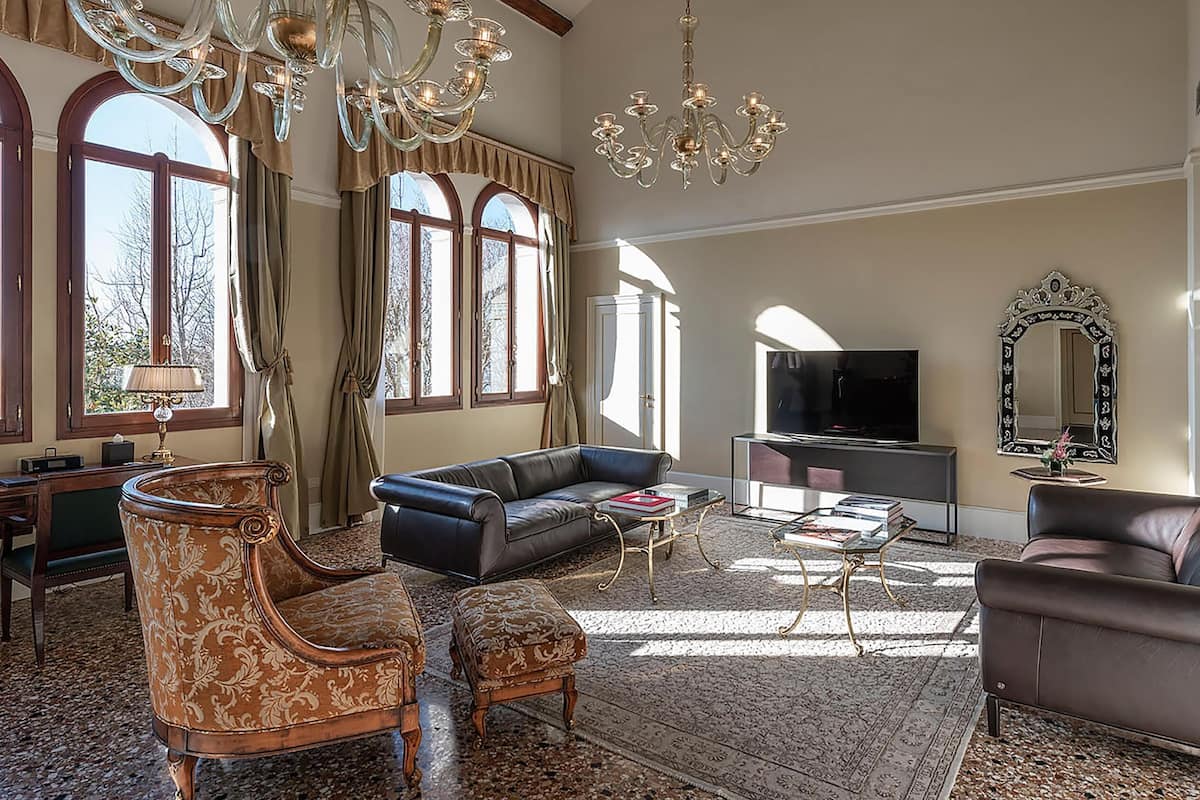 A spacious living room with a large sofa, a smaller armchair, a coffee table, and a television. There are large windows with curtains, a chandelier, and a mirror on the wall.