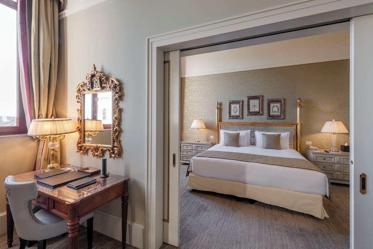 A hotel room with a large bed, two bedside tables with lamps, a wooden desk with a chair, and a mirror.
