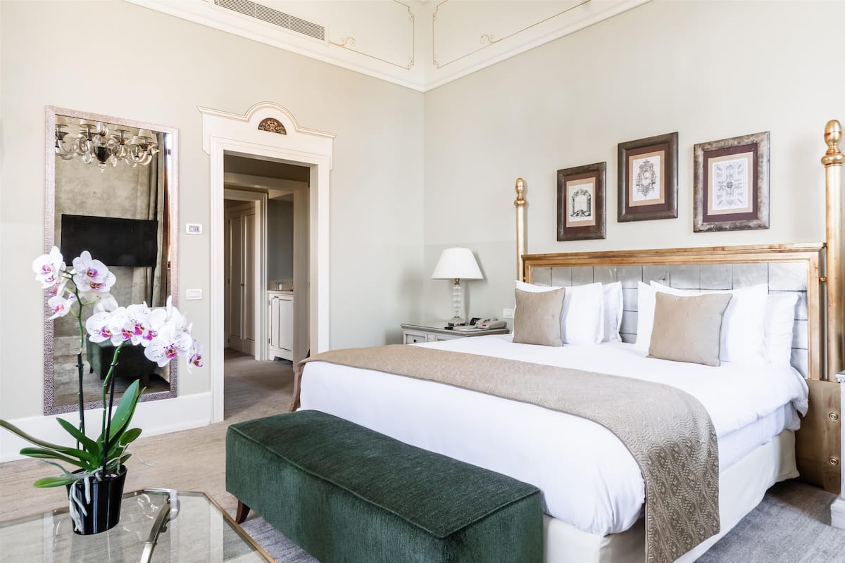 A hotel room with a large bed, a green bench, a glass table with a potted orchid, and a mirror with a chandelier.