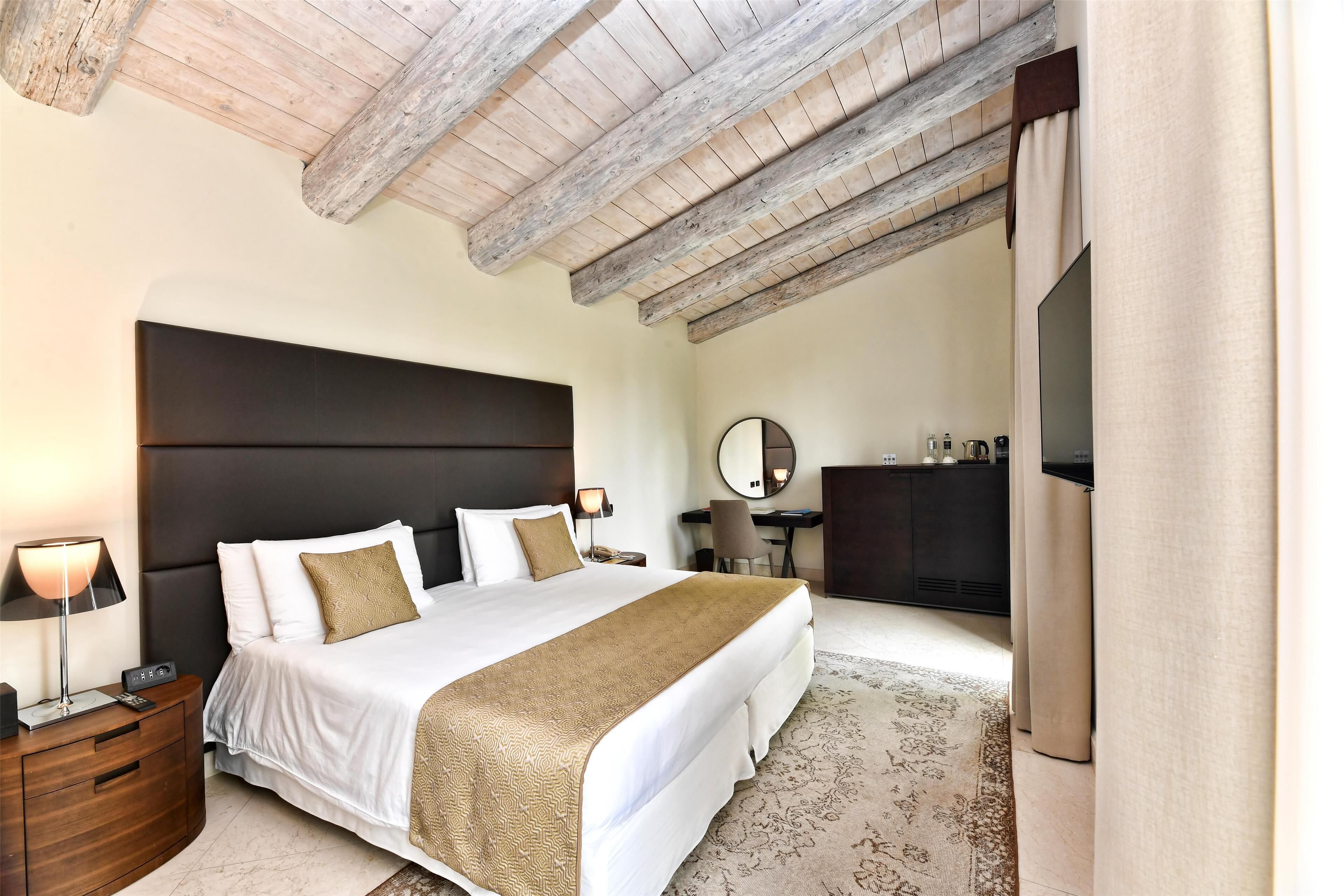 A bedroom with a large bed, a wooden ceiling, a desk, a chair, and a TV.