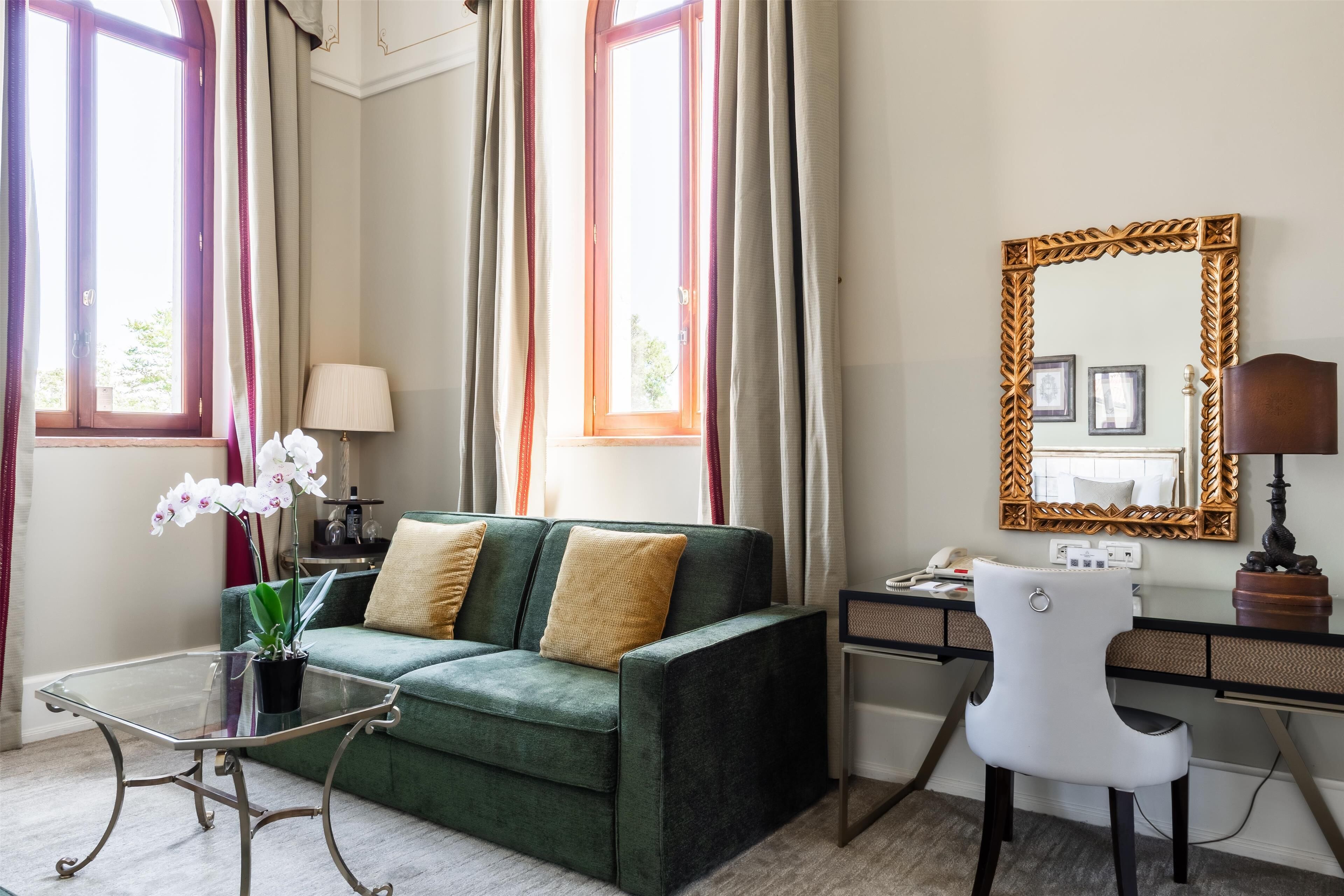 A room with a green sofa, a glass coffee table, a white chair, a desk, and a large mirror.
