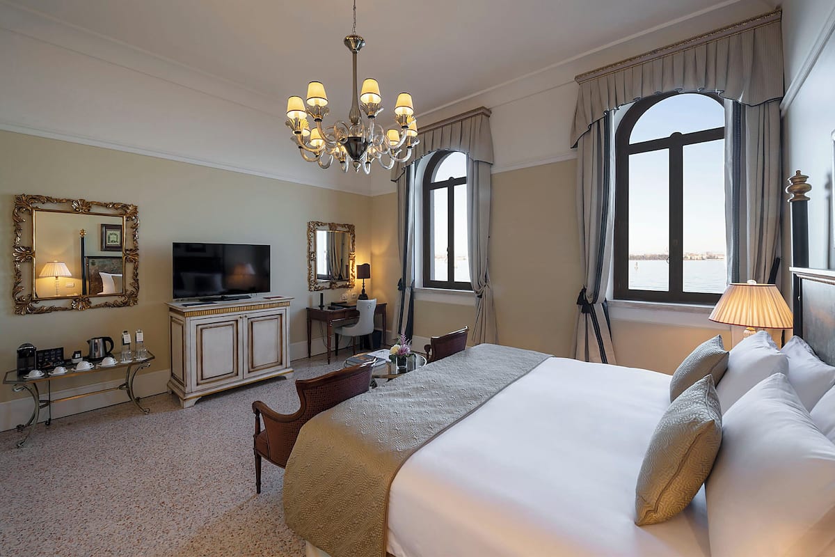 A hotel room with a large bed, a television, a desk with a chair, a mirror, and two windows with curtains.
