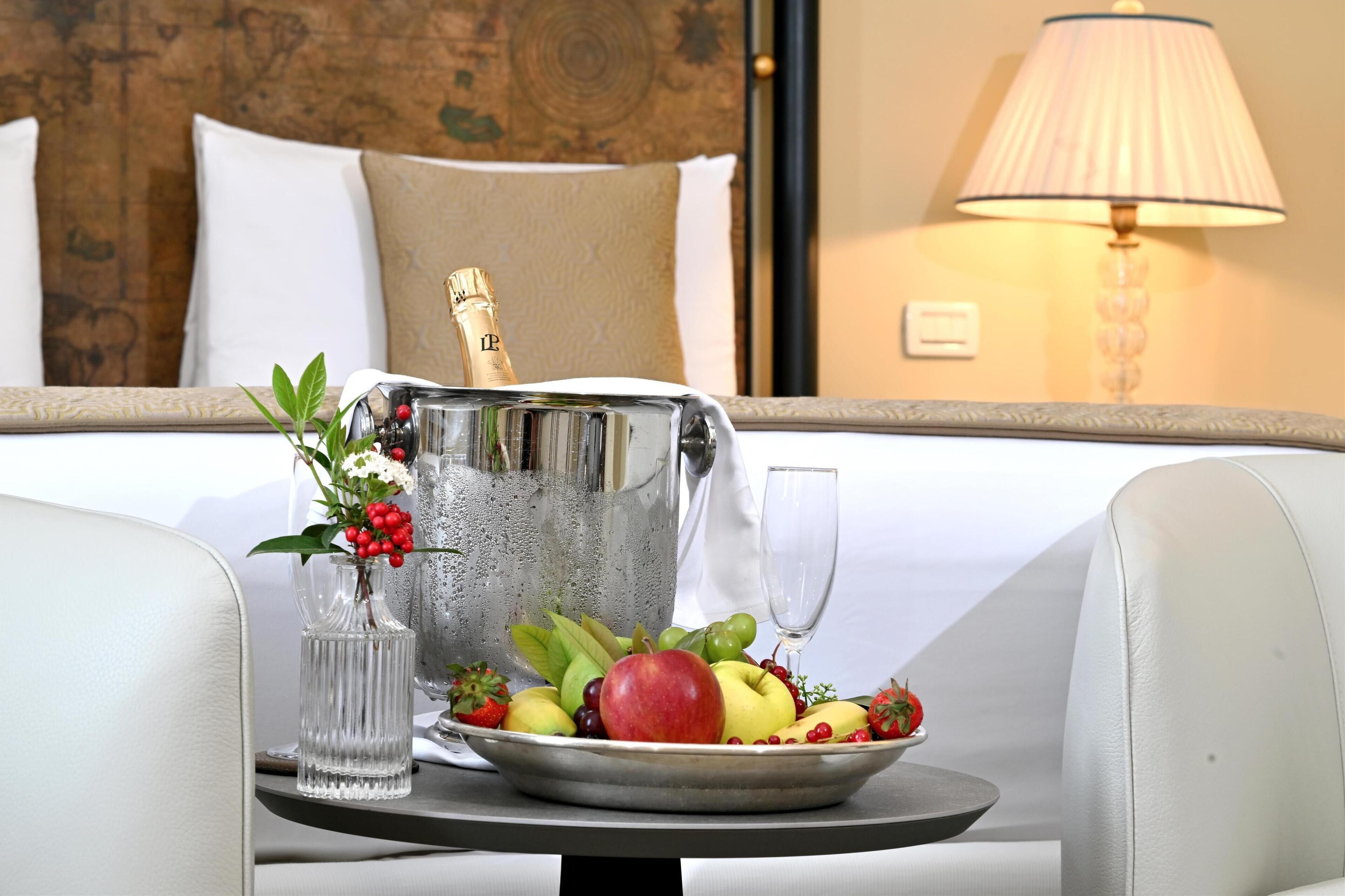 Habitación de hotel con una cama, una mesita de noche con frutero, una botella de champán y una copa.