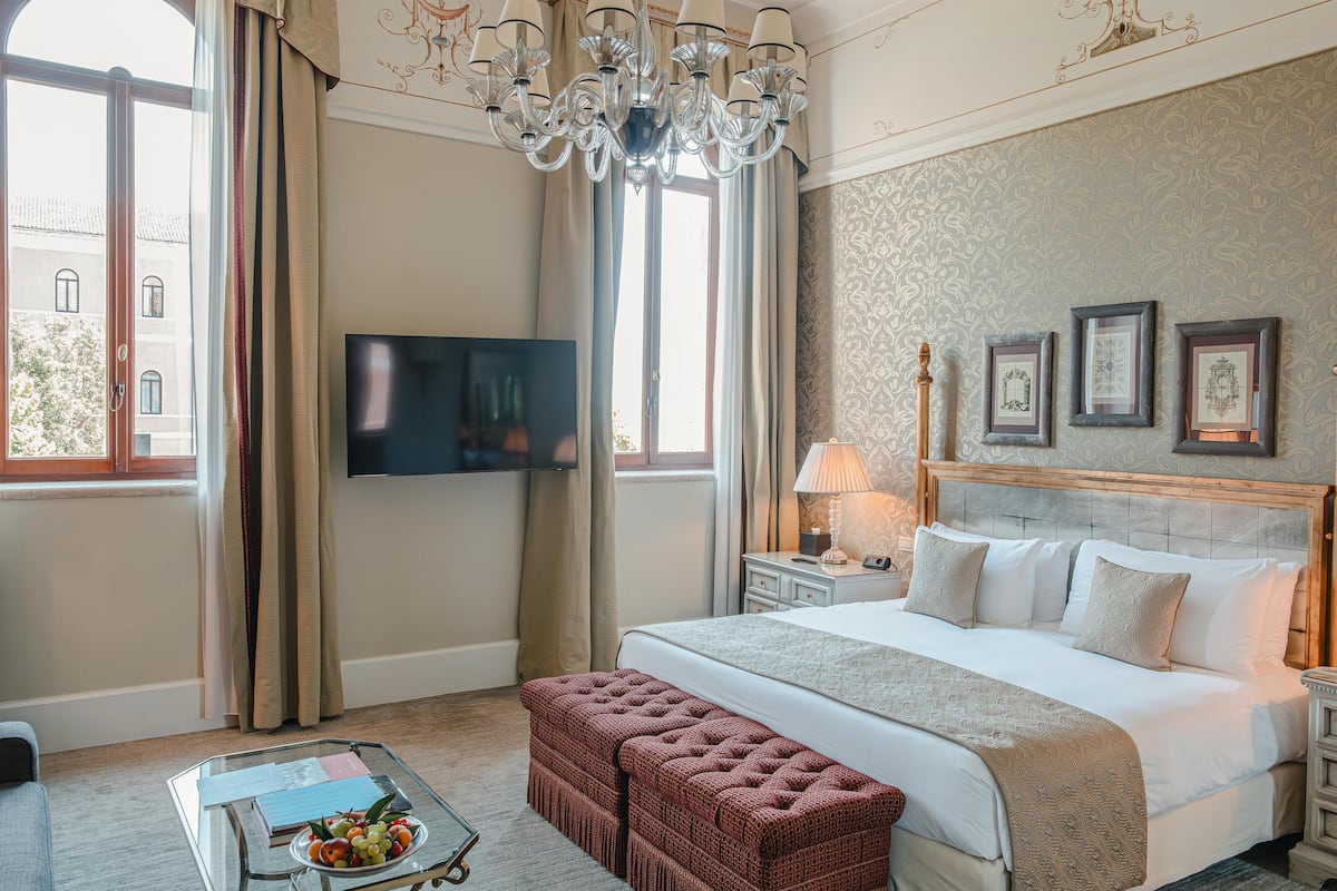 A hotel room with a large bed, a TV, a chandelier, and a view of the city through the window.