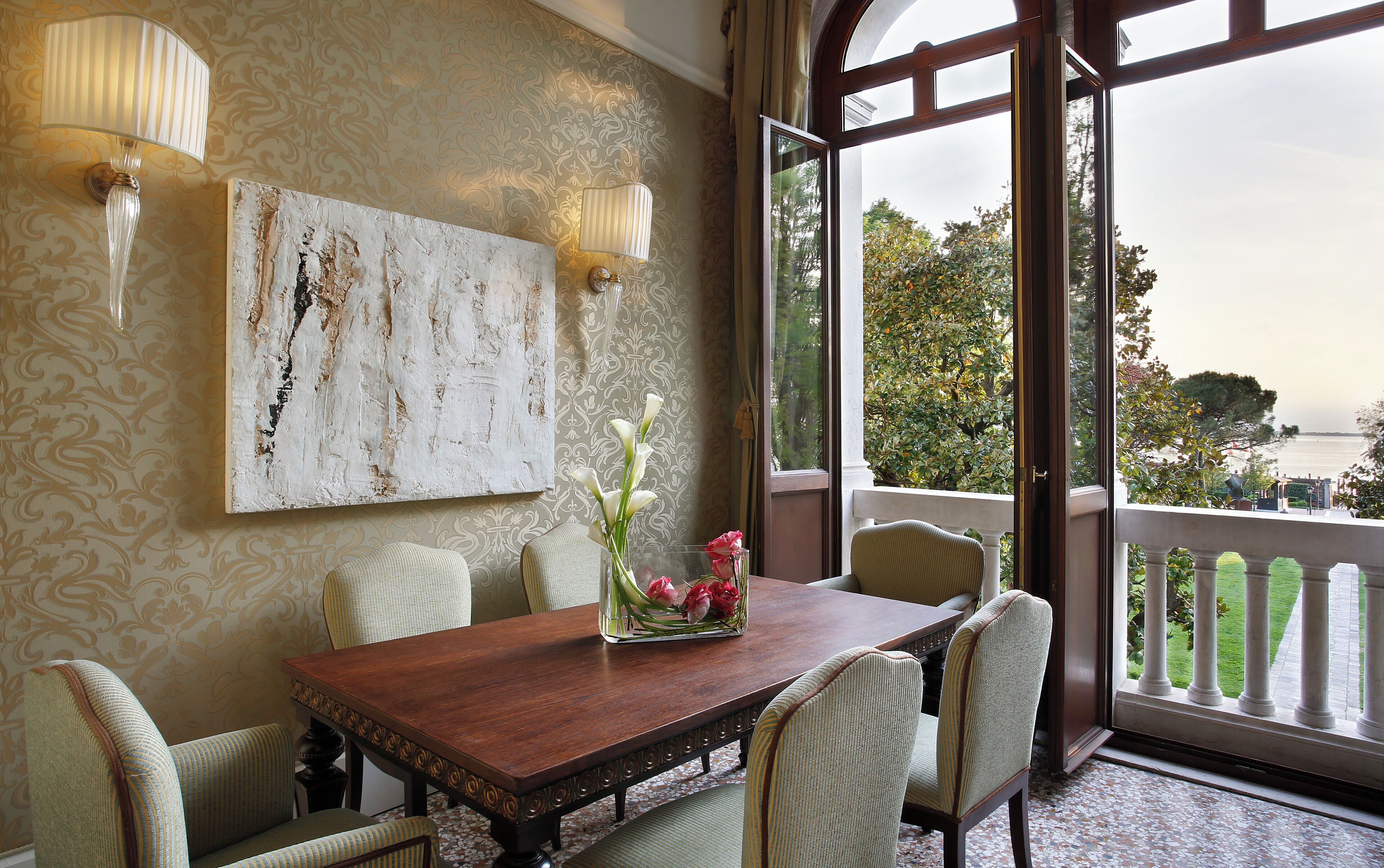 A dining area with a wooden table, six chairs, a vase with flowers, and a large window with a view of greenery and a body of water.