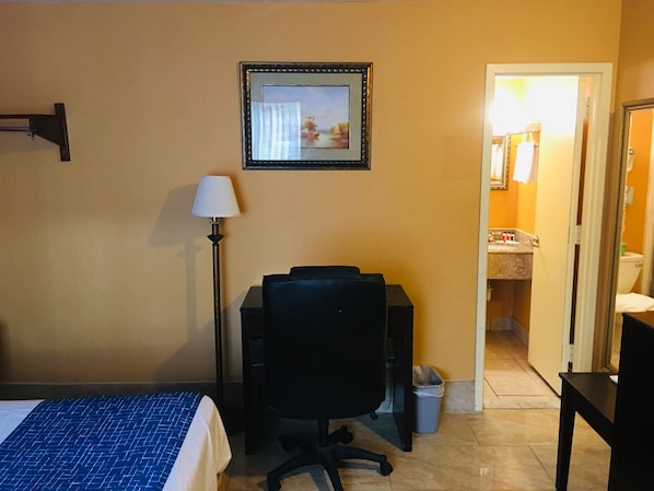A hotel room with a bed, desk, chair, lamp, and a framed picture on the wall.