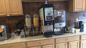 A kitchen counter with a waffle maker, a coffee machine, and two dispensers of cereal. - Travelodge by Wyndham Bay Shore Long Island (Bay Shore)