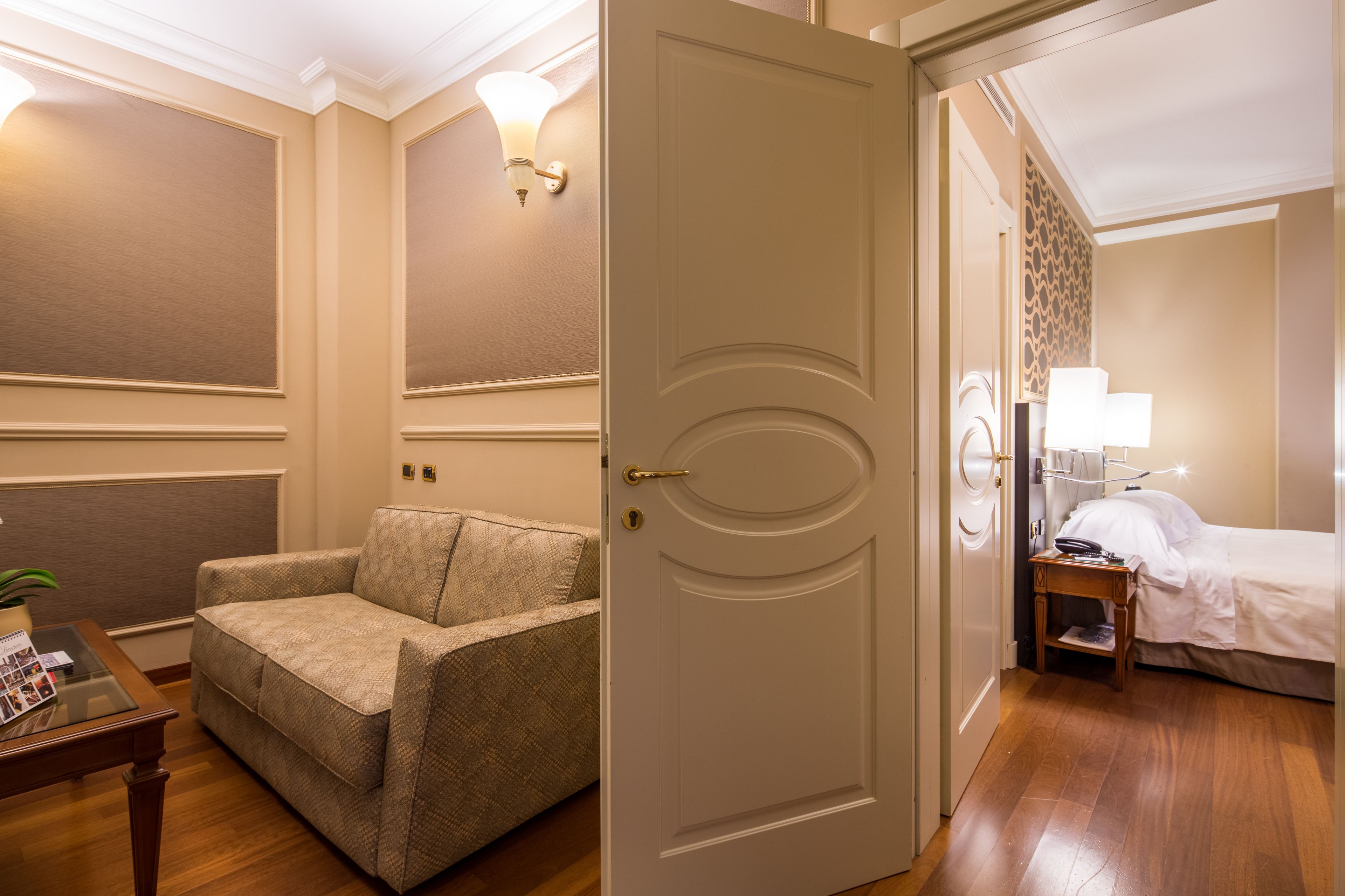 A hotel room with a beige sofa, a wooden coffee table with magazines, a bedside table with a lamp, and a bed with white linens.