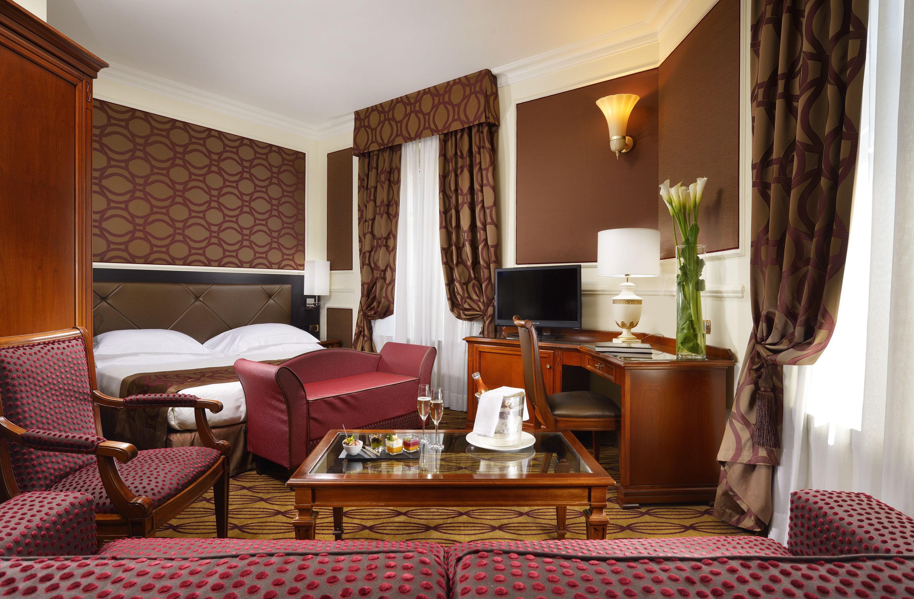 Habitación de hotel con cama, sofá rojo, mesa de comedor con jarra y vasos, televisor y escritorio con lámpara.