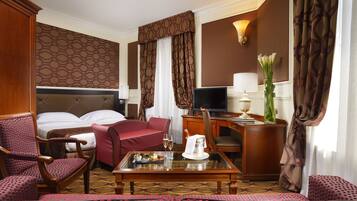 Habitación de hotel con cama, sofá rojo, mesa de comedor con jarra y vasos, televisor y escritorio con lámpara.
