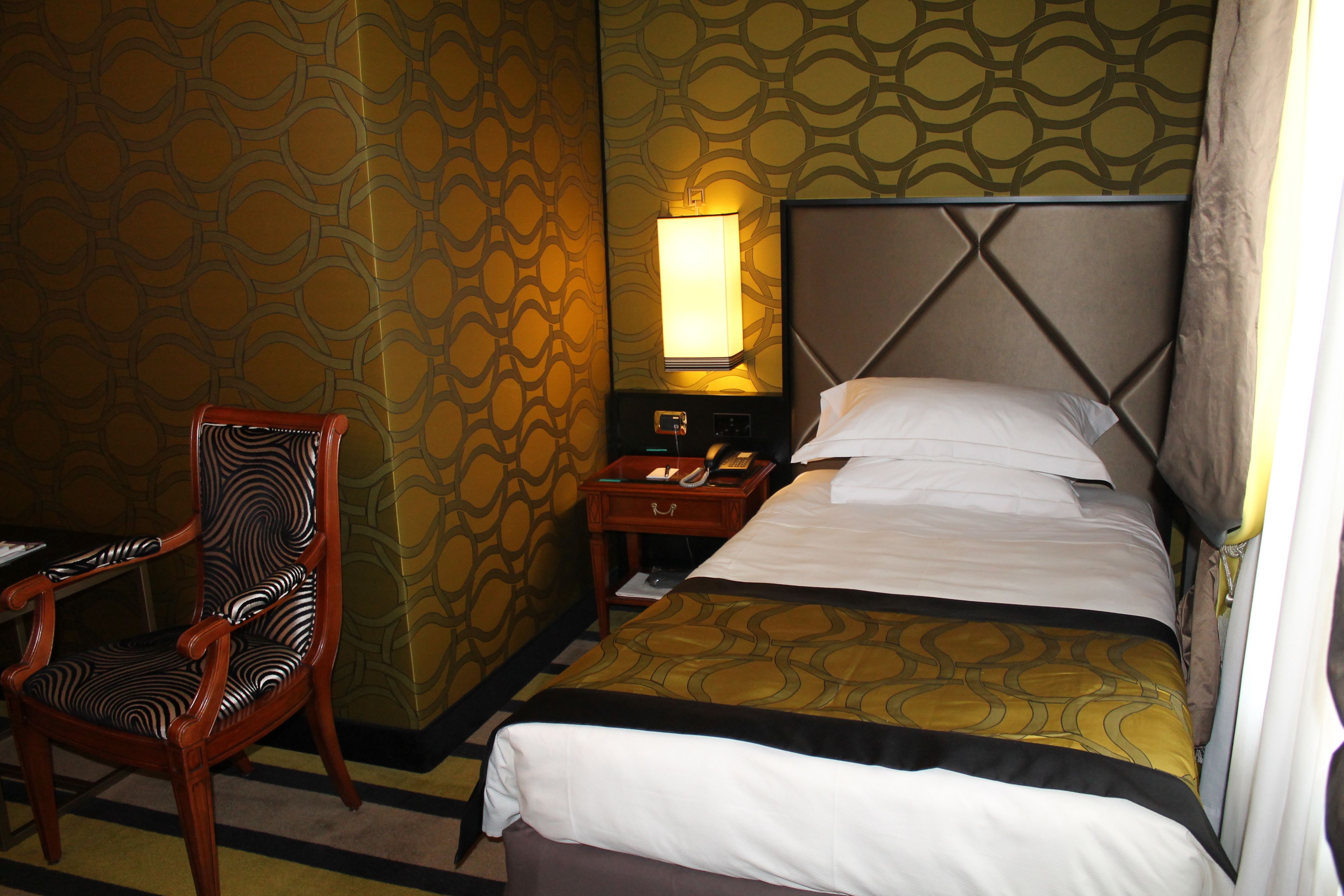 Ein Hotelzimmer mit Bett, Sessel, Nachttisch mit Lampe und Wandtapete mit Muster.
