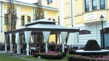 A gazebo with tables and chairs, surrounded by a well-manicured garden with hedges and flowers.