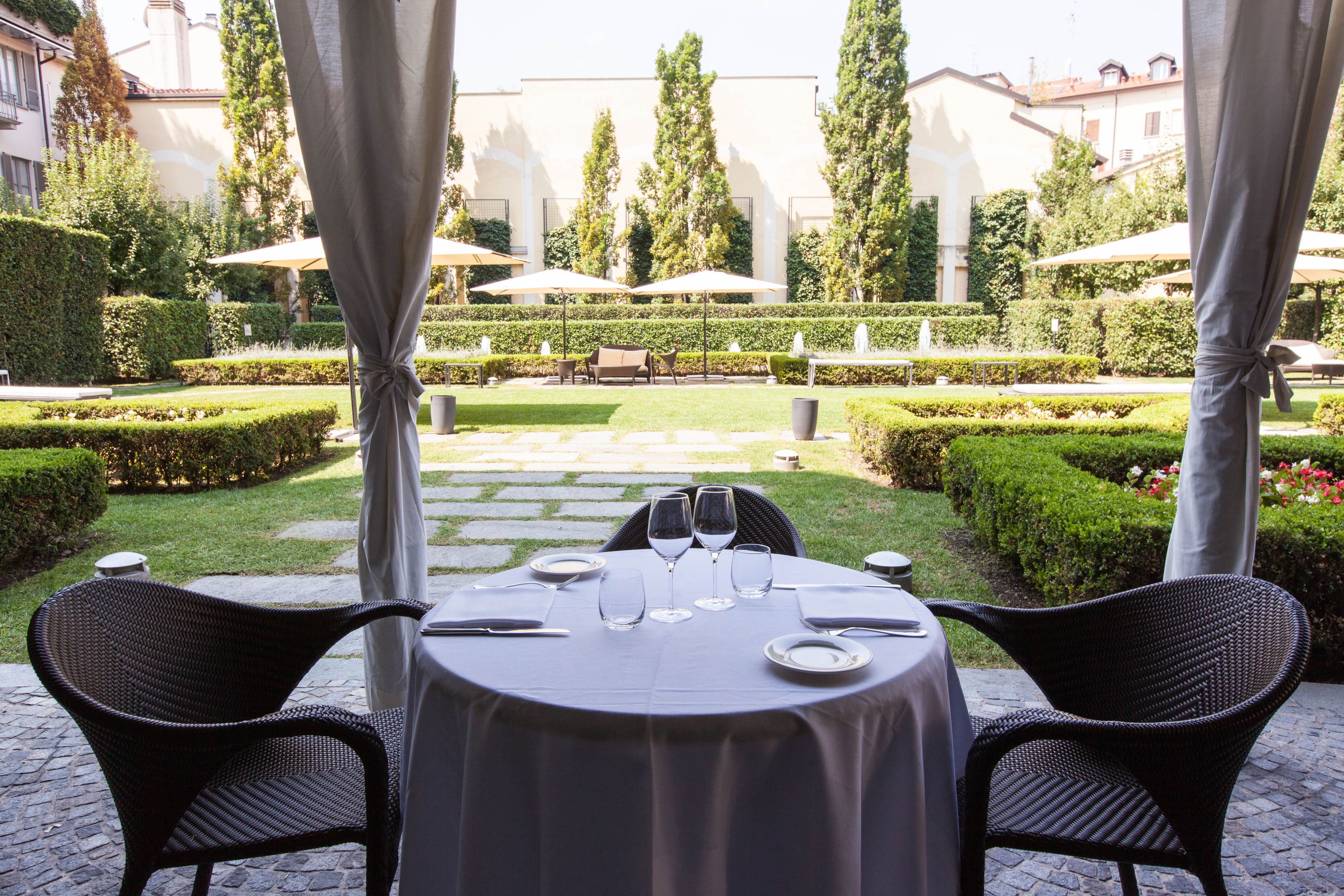 An outdoor dining area with a table set for two, surrounded by chairs, and a view of a well-manicured garden with hedges and trees.