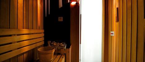Un sauna en bois, avec des bancs et un seau, une porte donnant sur l’extérieur et une petite étagère avec quelques objets.