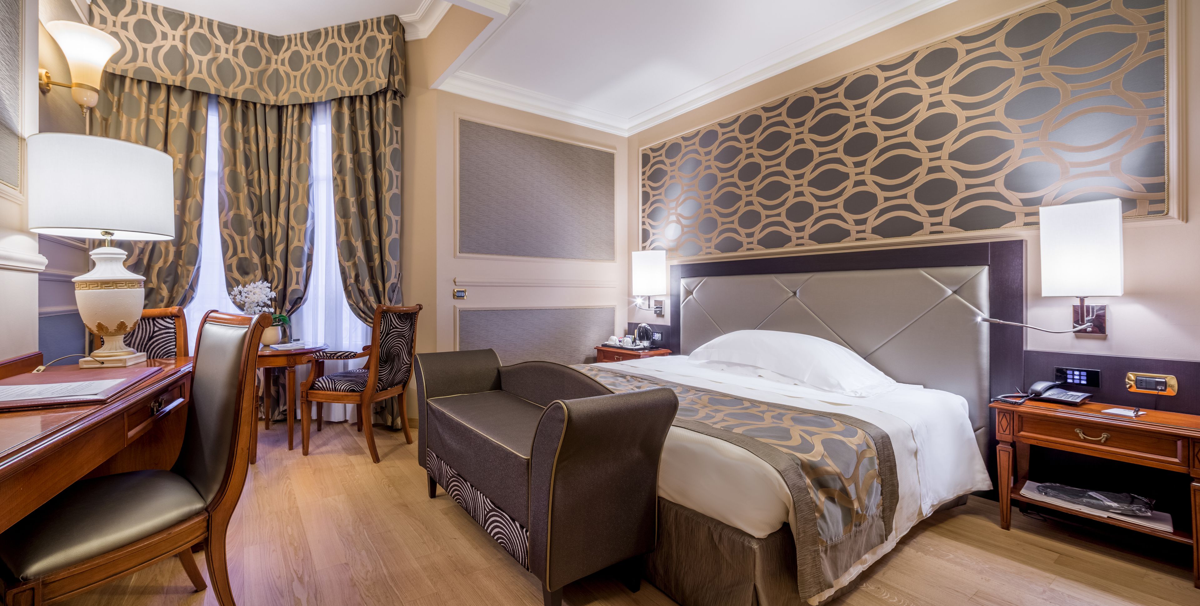 A hotel room with a large bed, a desk with a chair, a small table with a lamp, and a decorative wall panel.