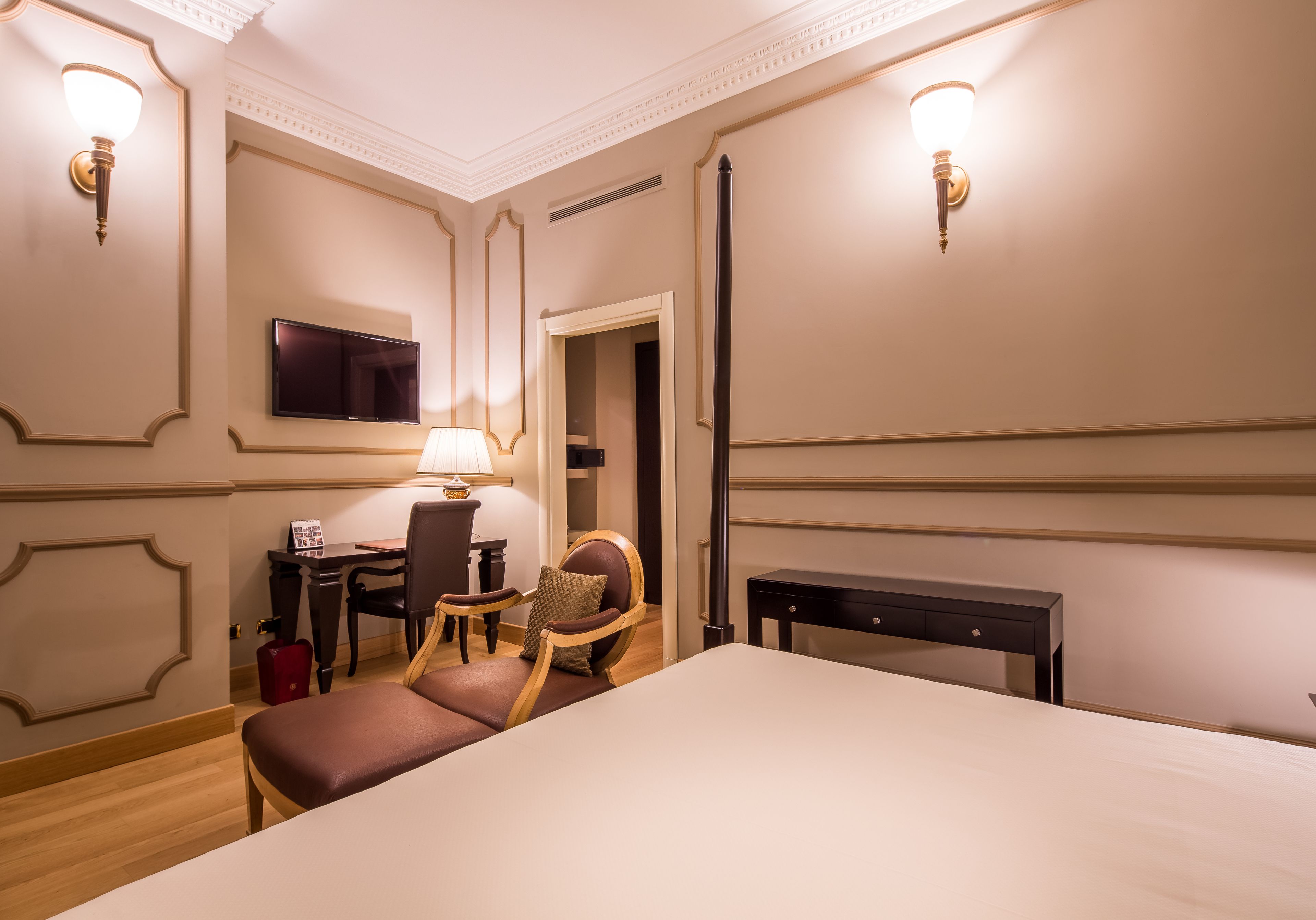 A hotel room with a large bed, a desk with a lamp, a chair, a television, and a wardrobe.
