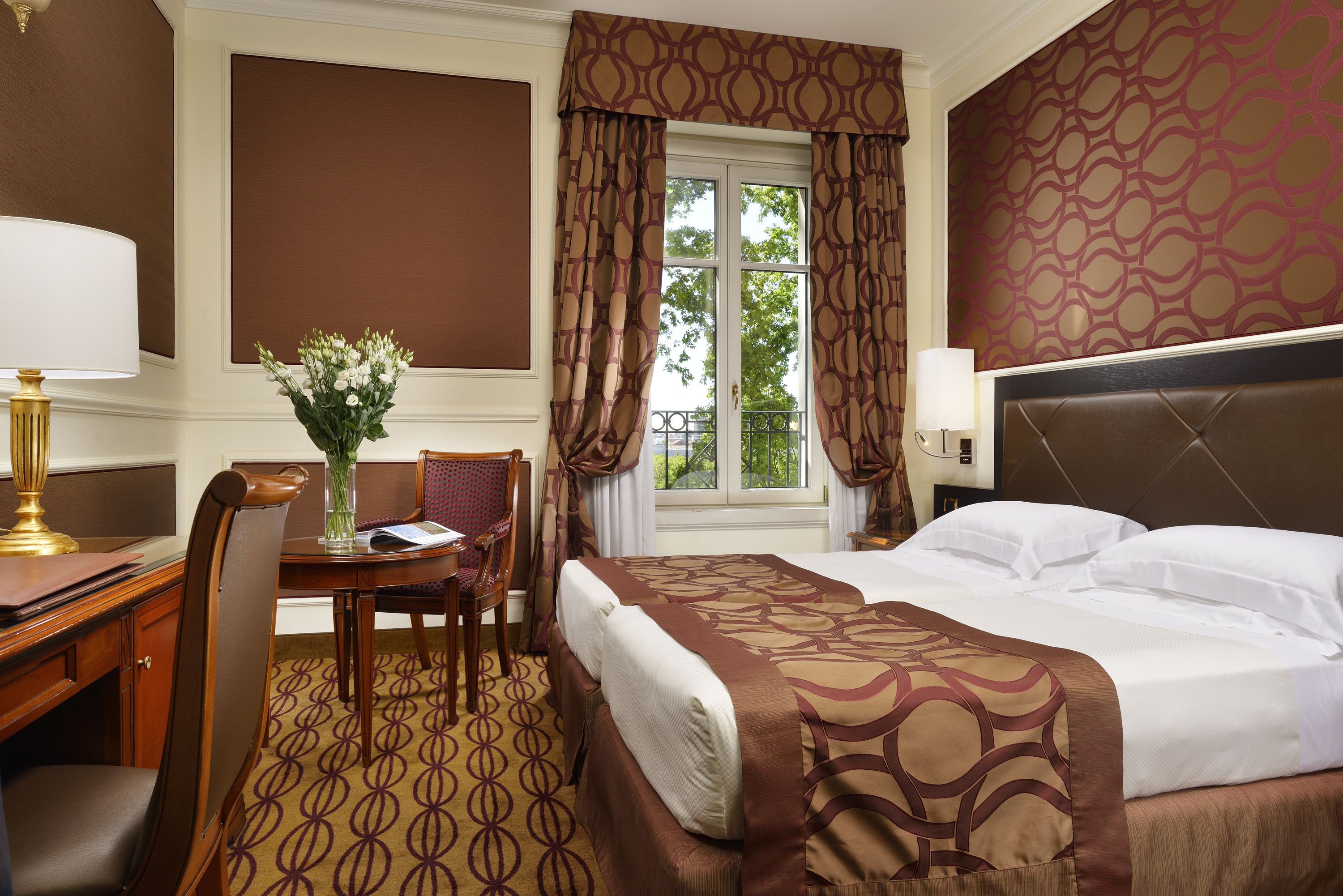 Een hotelkamer met een bed, een bureau, een stoel, een vaas met bloemen en een raam met gordijnen.