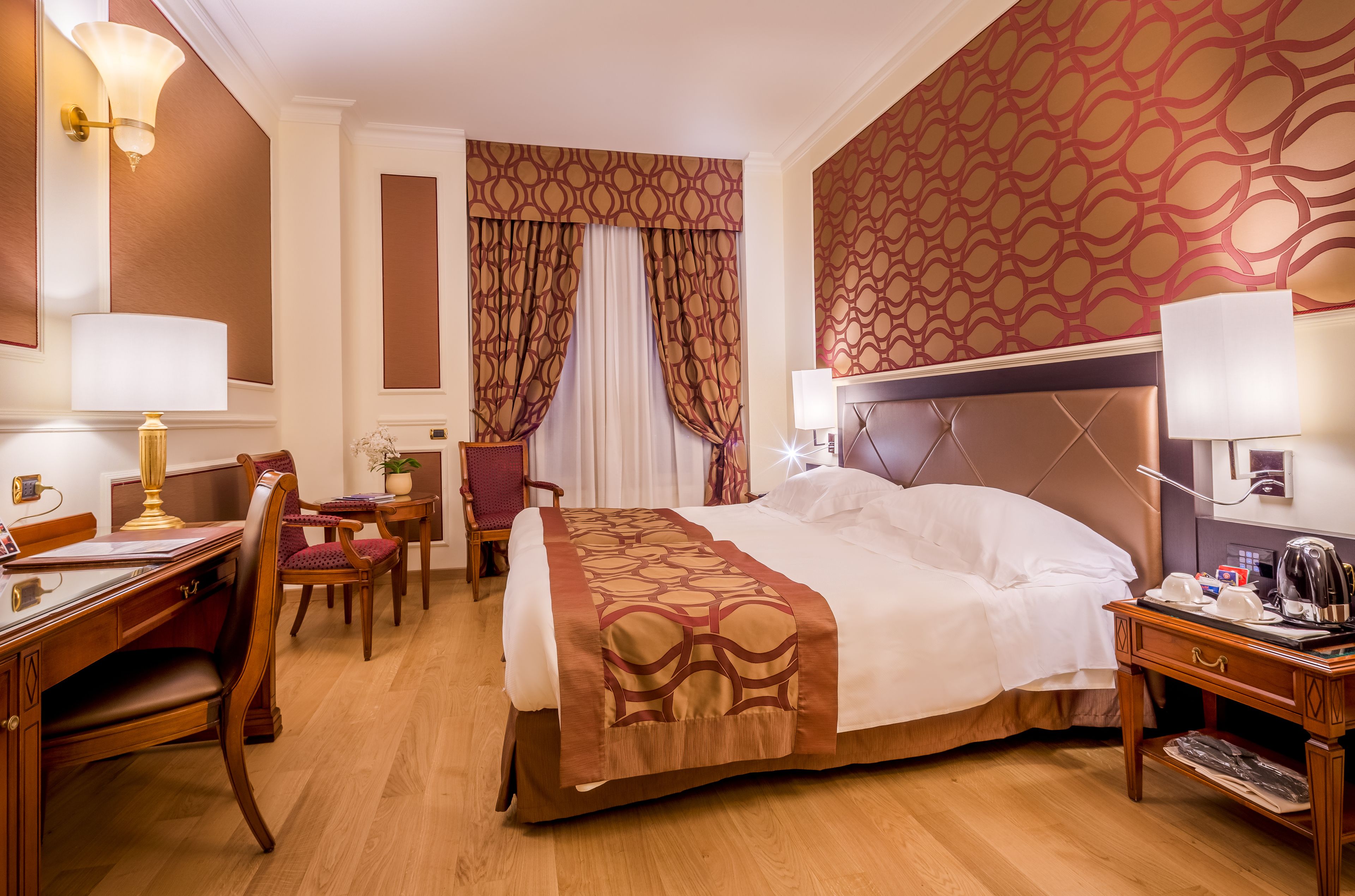 A hotel room with a large bed, a desk with a lamp, a chair, and a small table with a kettle.