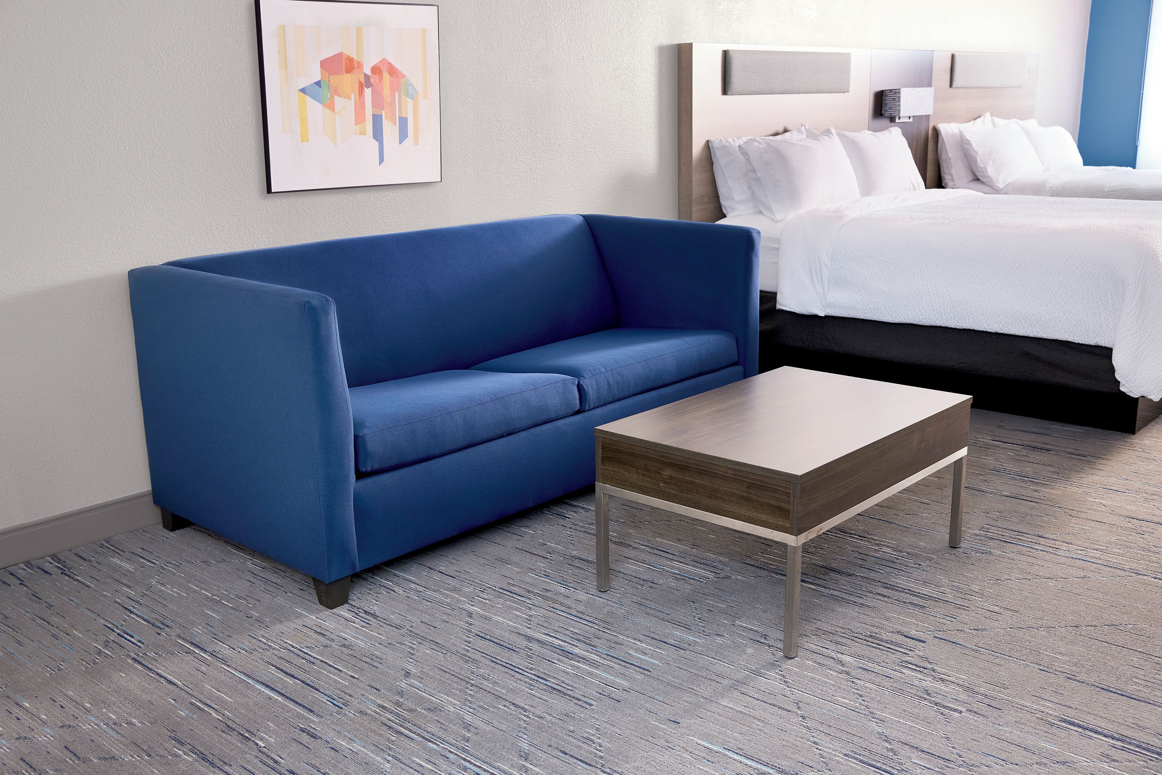A hotel room with a blue sofa, a wooden coffee table, and a bed with white bedding.