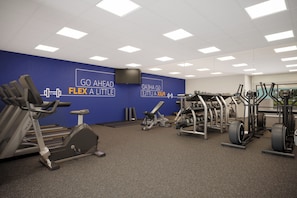 Un gimnasio con diversos equipos de ejercicio, una pared azul con texto motivacional y un televisor de pantalla plana montado en la pared.