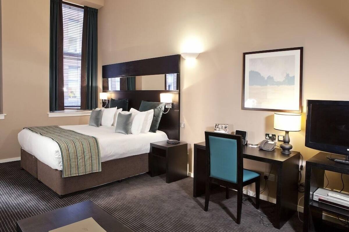 A hotel room with a bed, bedside tables, a desk with a chair, a TV, and a framed picture on the wall.