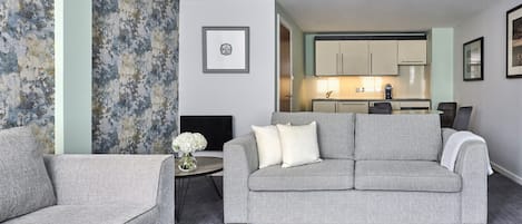 A living room with a grey sofa, a small side table with a vase, and a TV. There is a kitchen in the background with cabinets and a coffee machine.