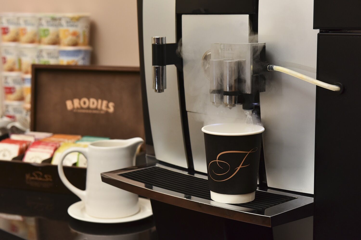 A coffee machine with a cup of coffee on a tray, a white pitcher, and a box labeled "BRODIES" in the background.