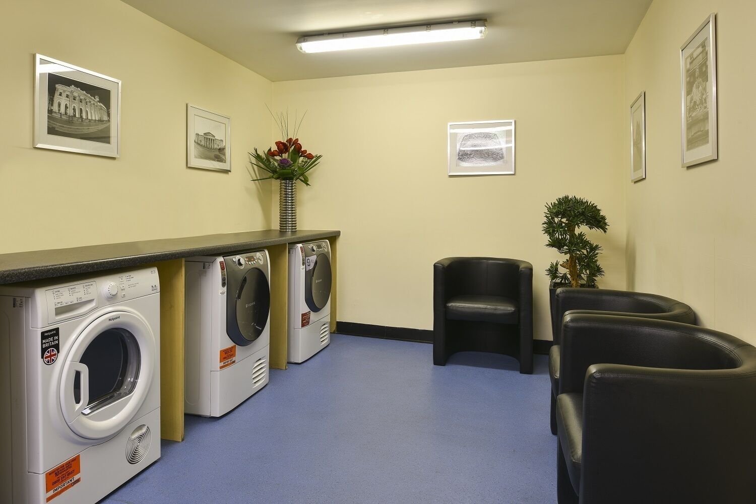 A laundry room with two washing machines, a vase with flowers, framed pictures on the wall, and black leather chairs.