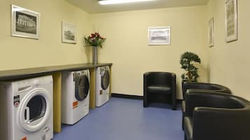 A laundry room with two washing machines, a vase with flowers, framed pictures on the wall, and black leather chairs.