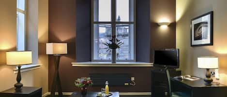 A hotel room with a sofa, a small table with a vase of flowers, a desk with a chair, a television, and a window with a view of buildings.