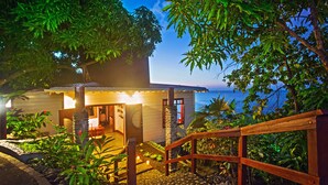 Een houten huis met een stenen trap naar een veranda, omringd door weelderige begroeiing en uitzicht op de oceaan.
