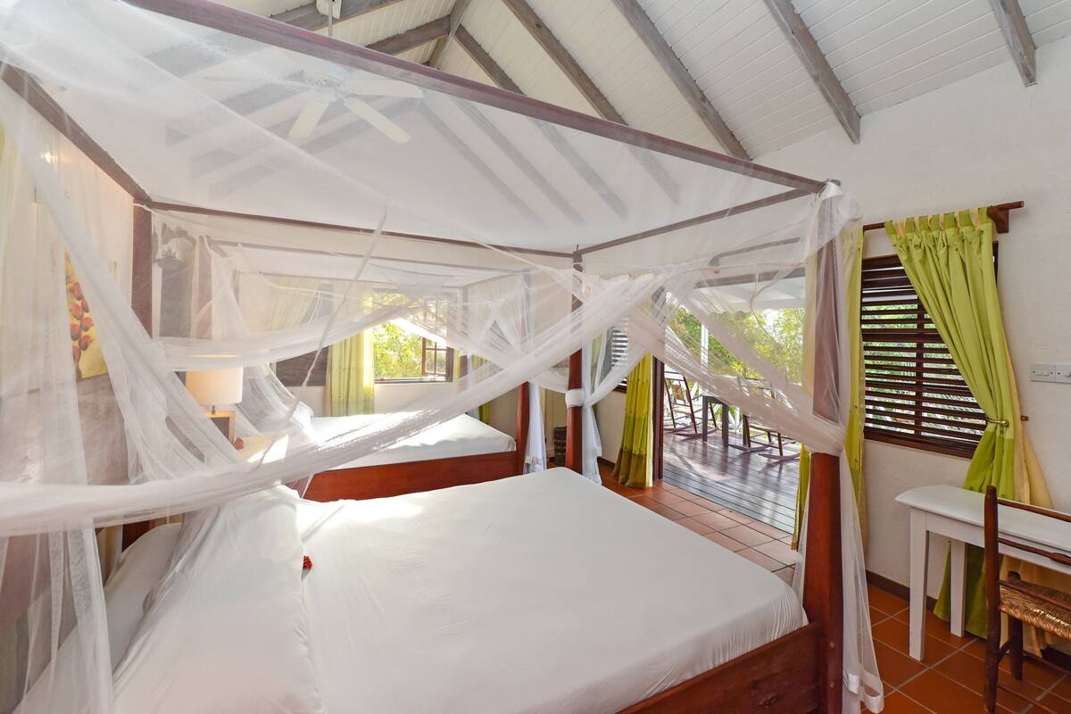 A room with a bed under a mosquito net, a window with curtains, and a small table.