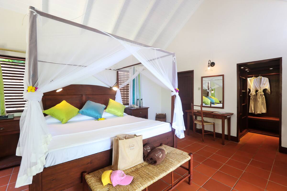 A bedroom with a canopy bed, colorful pillows, a bedside table, a mirror, a wooden bench, and a wardrobe.