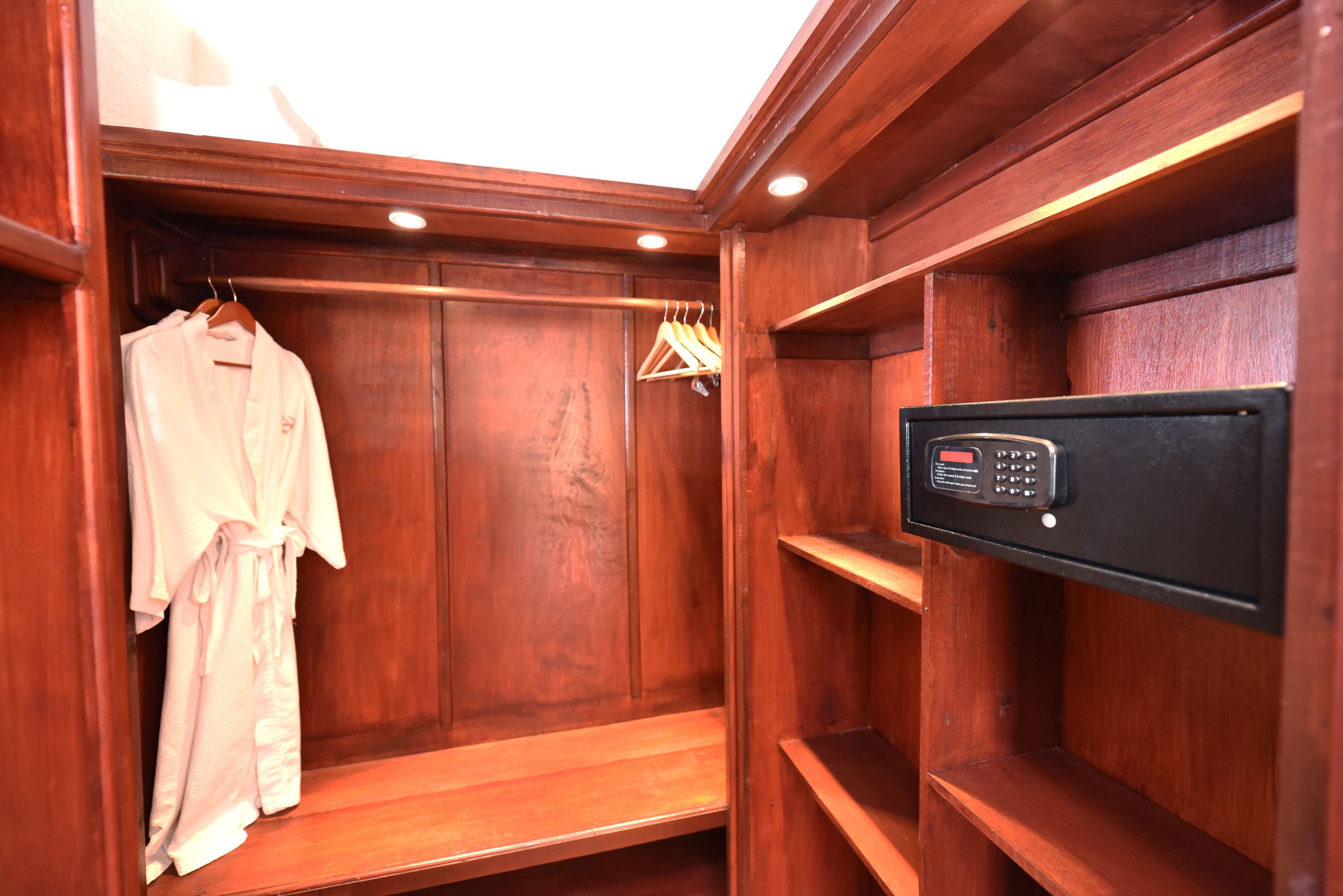 Un armario vestidor de madera con bata colgada, estantes de madera y una caja fuerte electrónica negra.