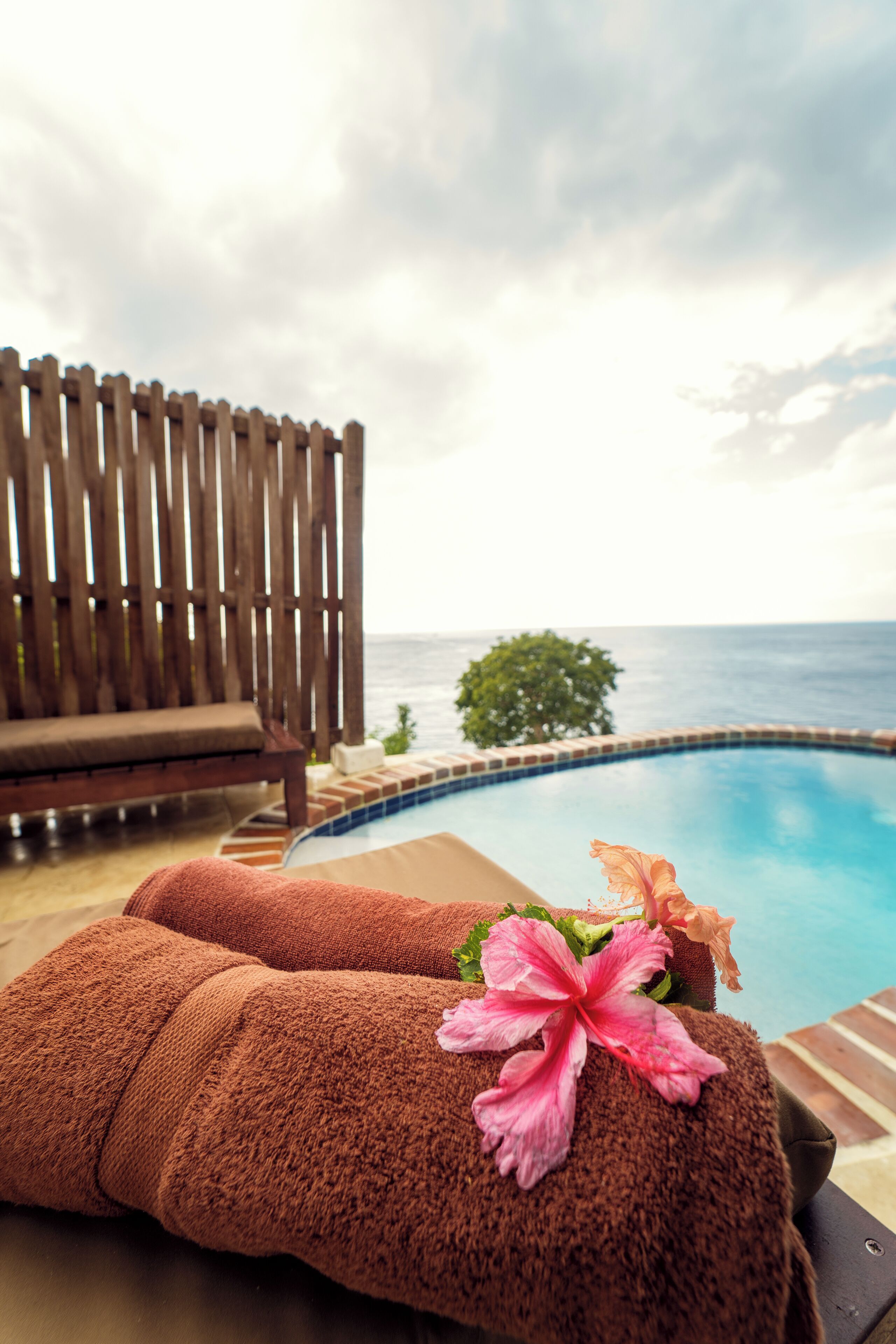 Zona junto a la piscina con valla de madera, una silla de descanso y toallas con estampado de hibisco.