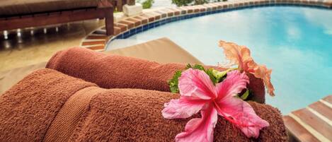 Zona junto a la piscina con valla de madera, una silla de descanso y toallas con estampado de hibisco.