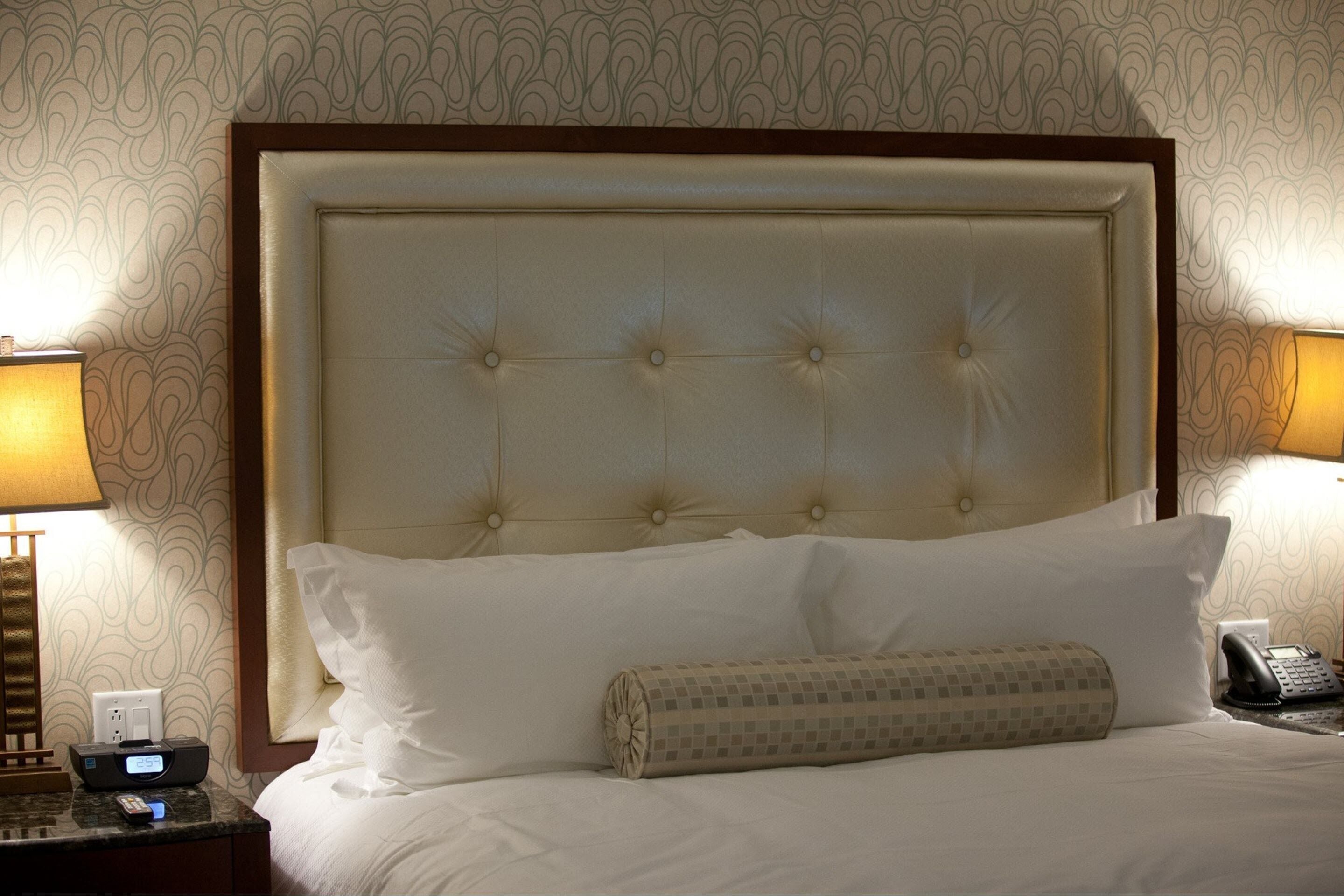 Hotelkamer met een gebobbelde hoofdbord, wit beddengoed, een gestreept kussen en een nachtkastje met een lamp, telefoon en digitale klok.