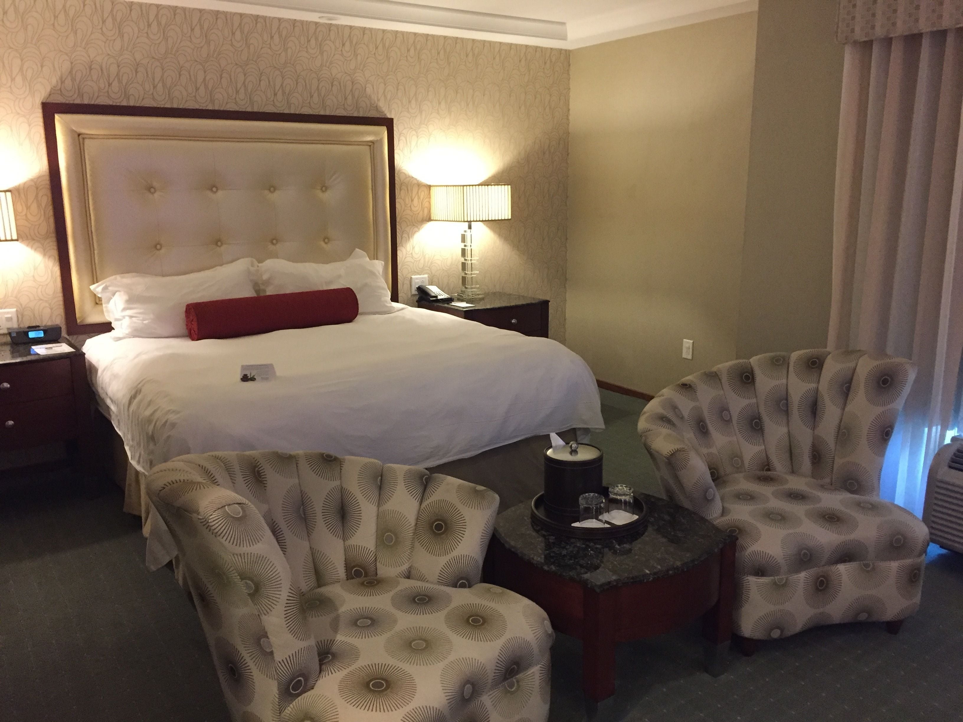 A hotel room with a large bed, two armchairs, a small table, and a nightstand with a lamp.