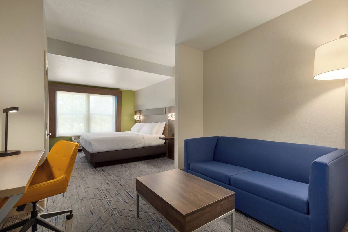 A hotel room with a bed, a blue sofa, a wooden coffee table, a yellow chair, and a desk.