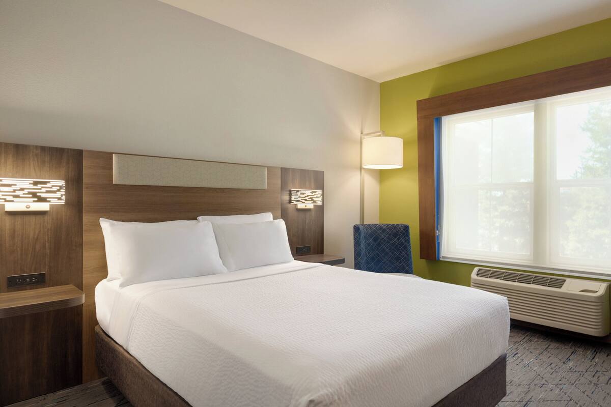 A hotel room with a large bed, a nightstand, a wall-mounted lamp, and a window with a view.