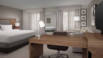 Habitació d'hotel moderna amb un llit gran, un escriptori de fusta, una cadira, un sofà, una televisió i una finestra amb cortines.