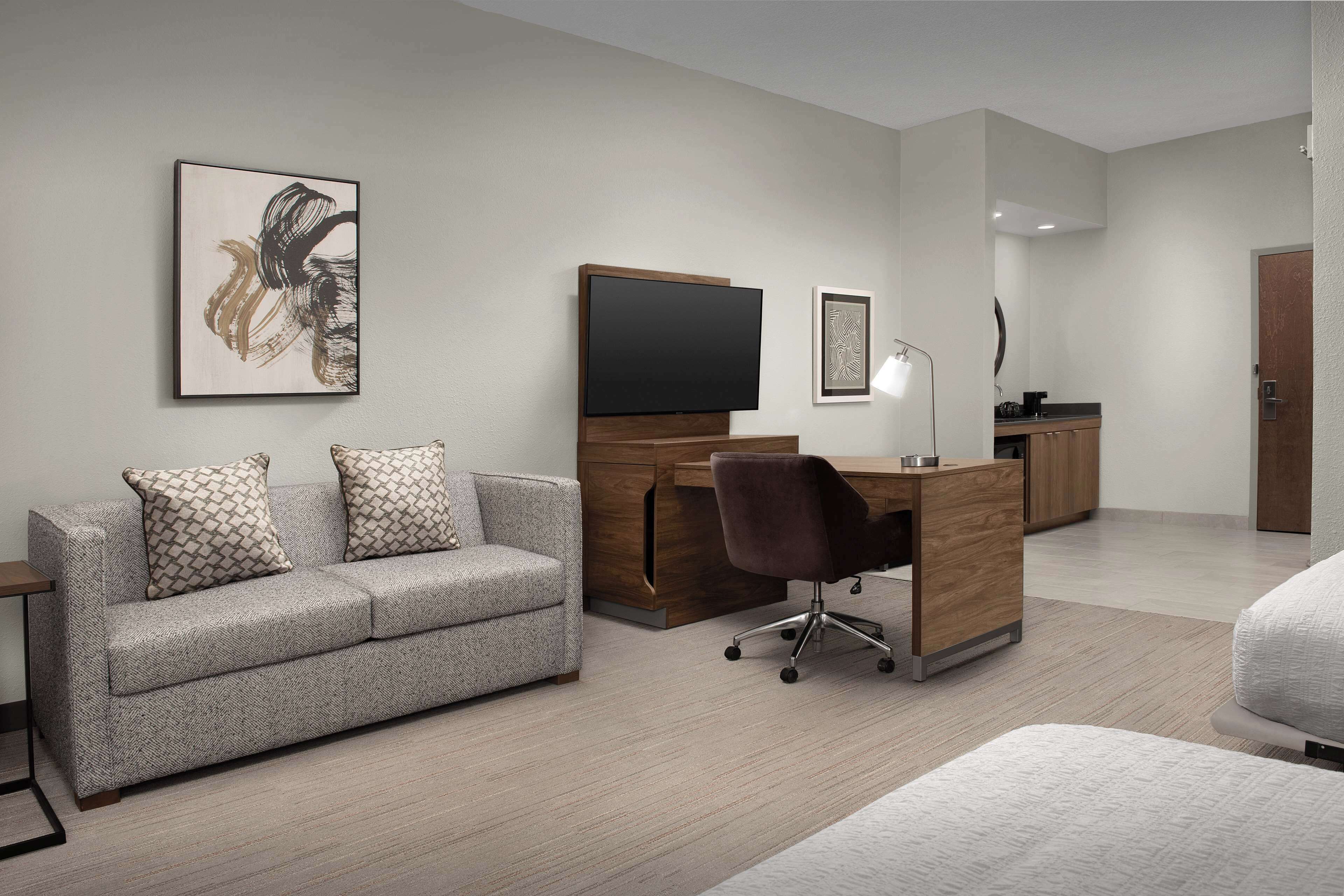 A modern hotel room with a sofa, a desk, a chair, a flat-screen TV, and a mirror.
