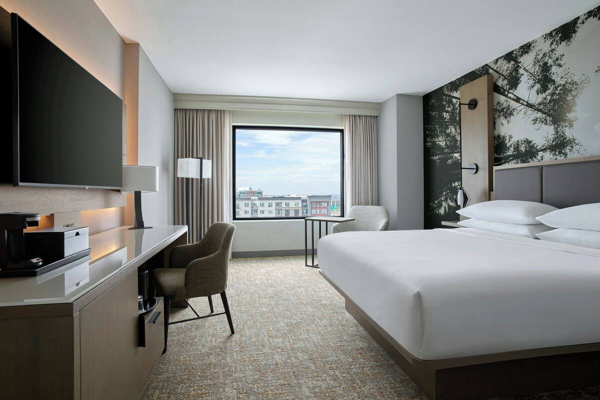 Habitación de hotel con cama, escritorio con computadora, silla, televisor y ventana con vista a la ciudad.